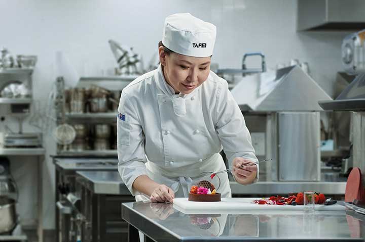 A young pastry chef student carefully adding finishing touches to a dessert in a commercial kitchen.