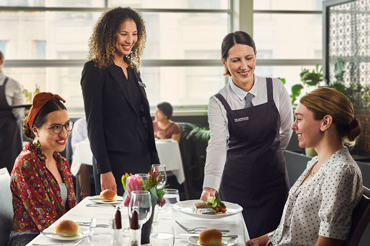 Trainee server presenting dish to guests at restaurant table under the supervision of her female teacher.