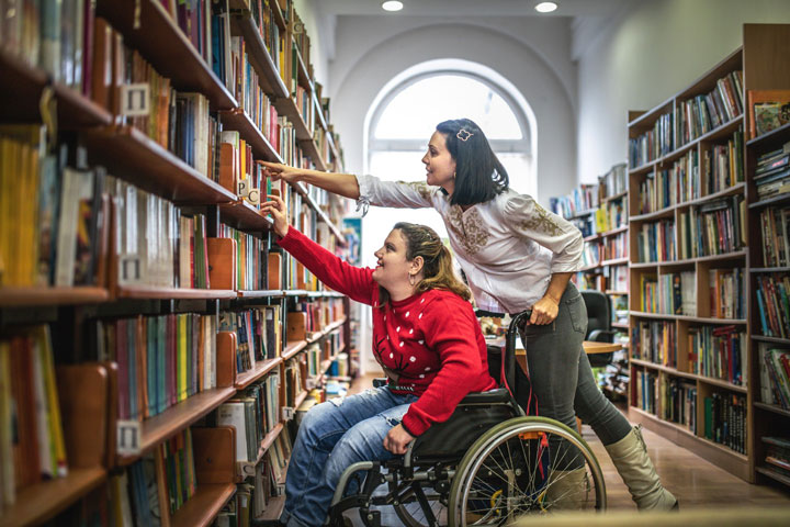 A woman in a wheelchair is reaching for a book on a high shelf in a library, while another woman beside her assists by pointing to a specific book. They are surrounded by rows of colourful bookshelves under soft lighting.