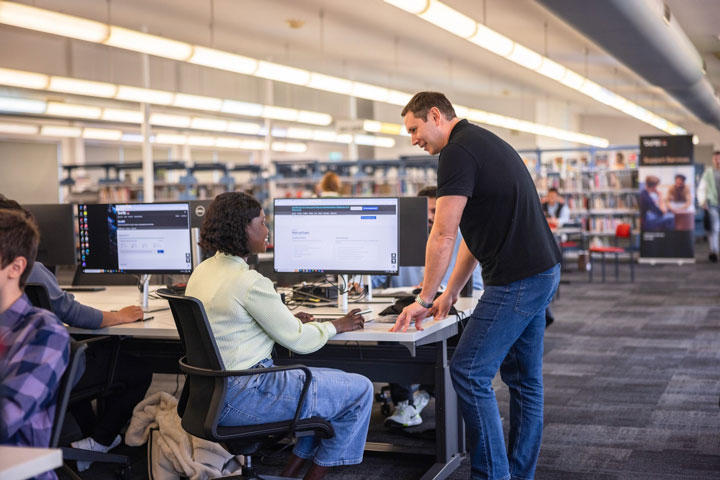 A man is helping a woman at the library. She is seated at a desktop computer and he is standing next to her. They are smiling and engaged in conversation. There are other students working in the background.