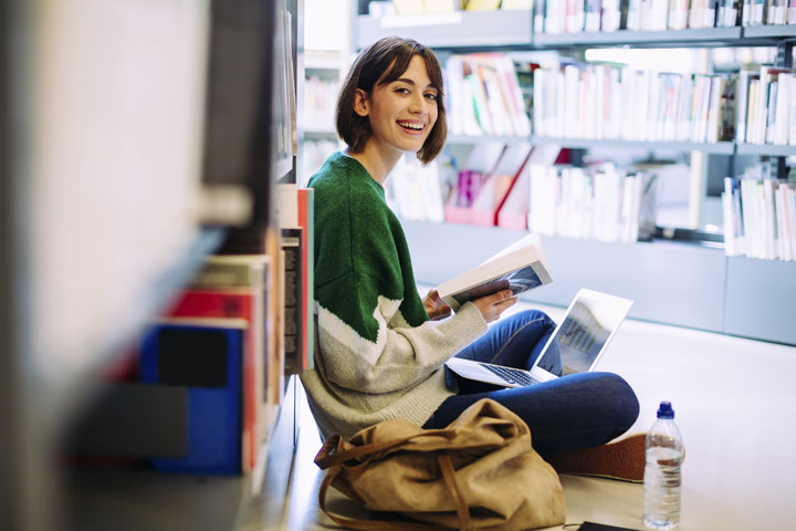 A woman with short hair sits on the floor of a library. She smiles at the camera as she sits with her laptop with a book in hand.