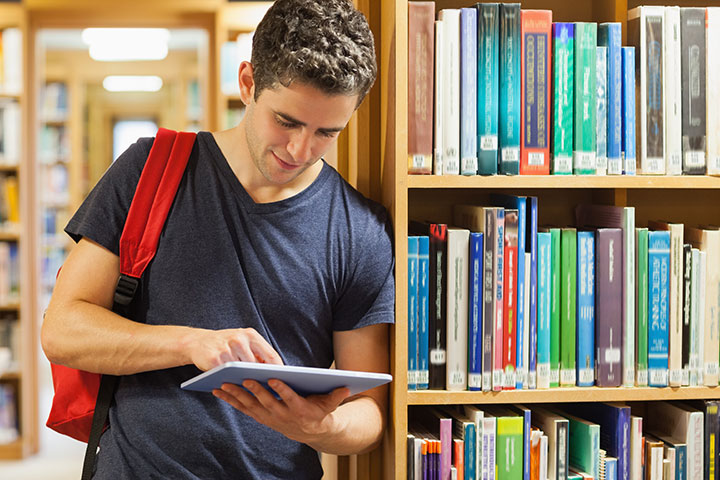 A man stands against a bookshelf in a library working on his touch-screen device. He has a backpack on and is working on his task.