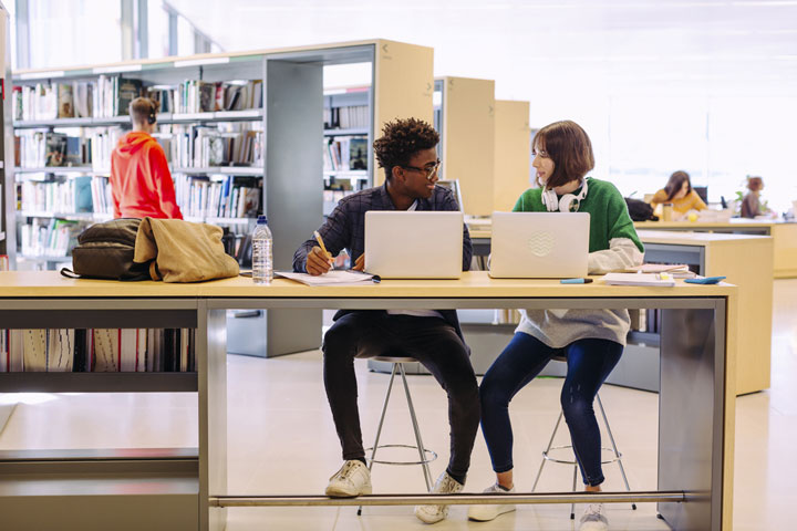 Two students sit in the library with laptops in front of them, reviewing ɫֱpolicies and smiling at each other.