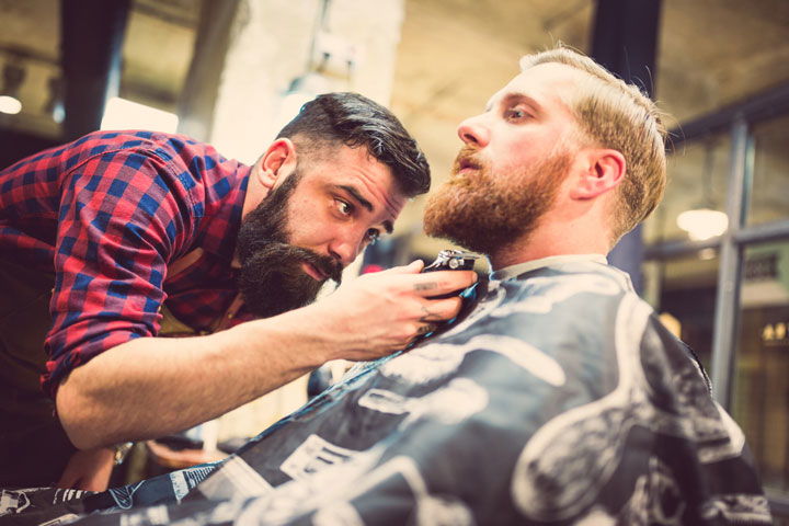 A barber in training, wearing a plaid shirt and apron, carefully trims the beard of a client seated in a barber's chair in a stylish barbershop setting. His focus clearly showcases his attention to detail and passion for the trade.
