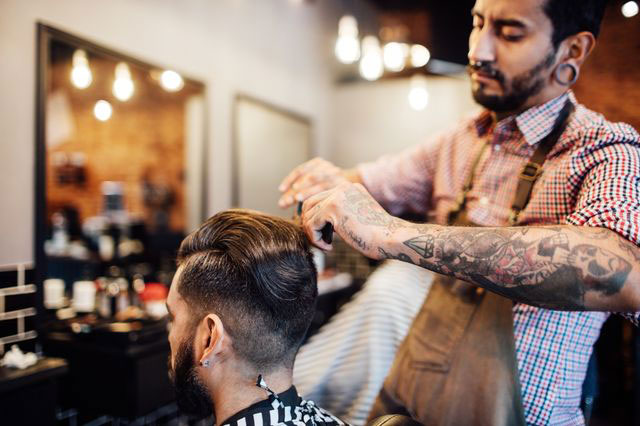 A barber with tattoos carefully trims a male client's hair in a modern barbershop. The client is seated in a striped cape, and the barbershop features mirrors, grooming tools, and exposed lightbulbs in the background