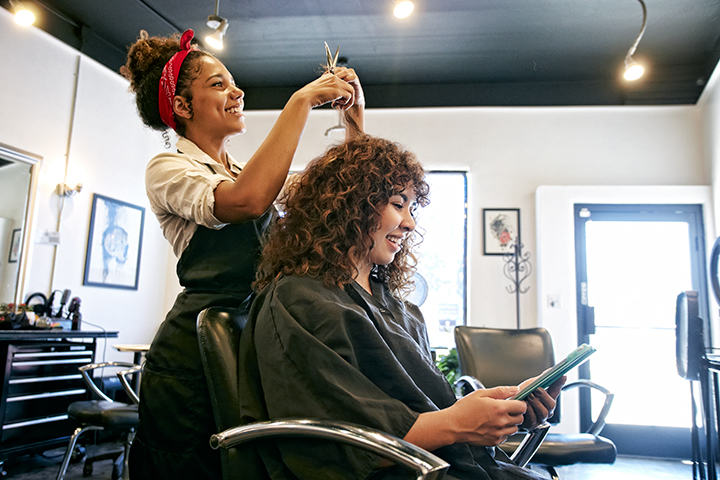 A young woman is wearing a red bandana in her hair and an apron. She is cutting another woman’s hair. The woman getting her curly hair cut, sits in smock reading. 