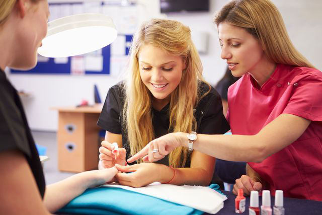 A young woman applies nail polish to a client's hand while an instructor in a pink uniform points to the nail to guide her. Nail polish bottles and a lamp are on the table.