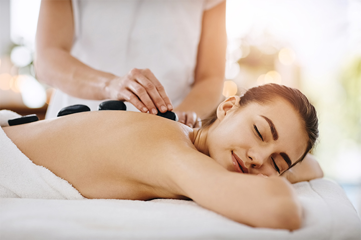 A woman lies face down on a massage table with a relaxed expression, receiving a hot stone massage. A massage therapist’s hands are adjusting smooth black stones on her back.