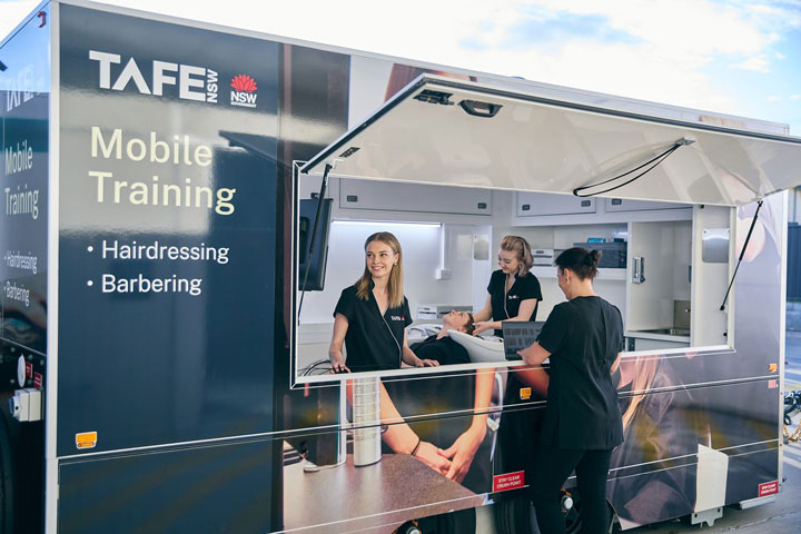 A TAFE NSW mobile training unit (MTU) featuring branded signage, where three hairdressing students collaborate. One student is washing a client's hair in the space.