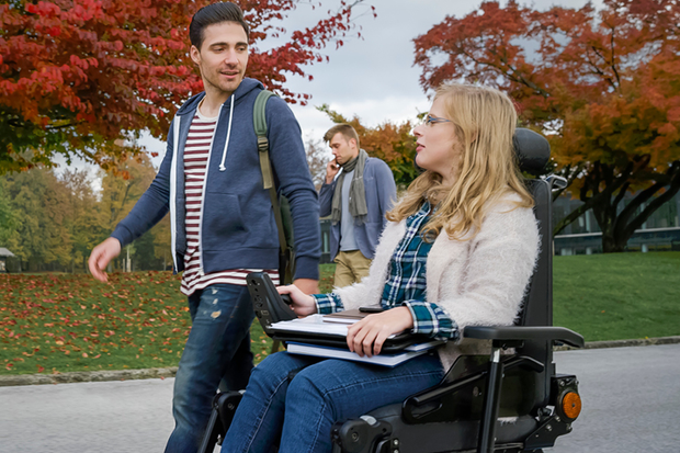 Student colleagues conversing and together on a park path, the young man walking and the young woman in a motorised wheel chair.