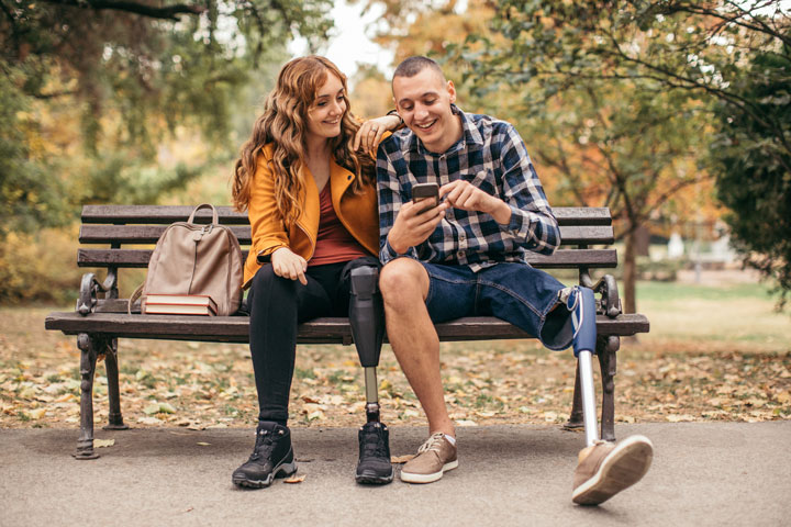 A young woman and young man, both with an artificial leg, sit closely. She has one arm resting on his shoulder while they both laugh, looking at something on his phone screen.