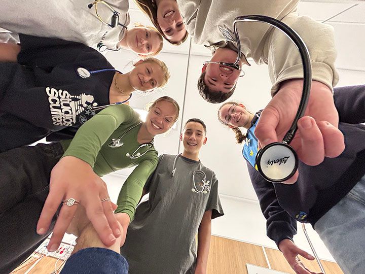 Group of young students with stethoscopes around their necks stand in a circle looking down at the camera. 