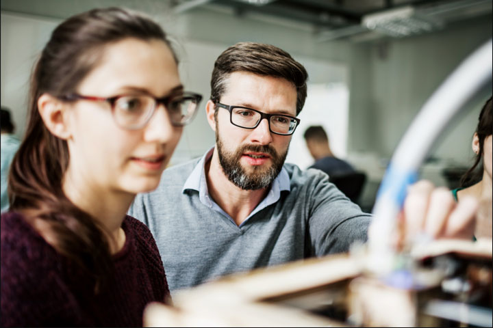 A male teacher guides a female student in a practical activity. Both wear spectacles.