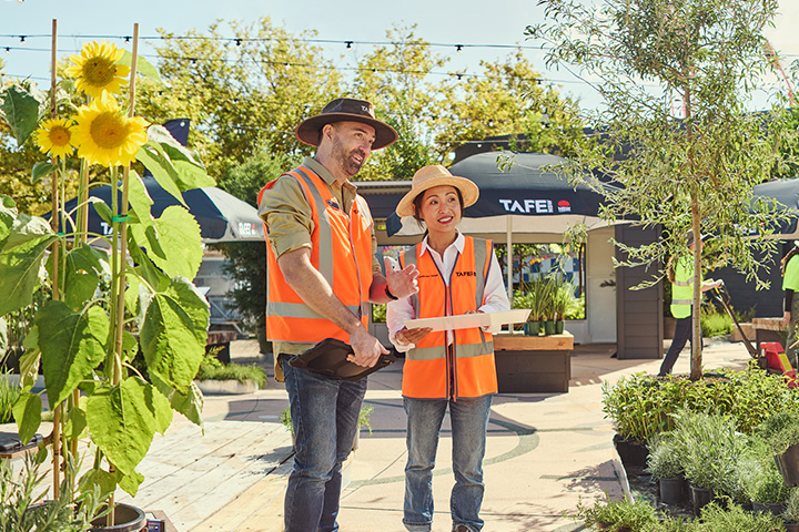 A horticulture student kneels beside a sunflower plant in a vibrant outdoor nursery, examining its leaves while their teacher stands nearby, offering guidance.