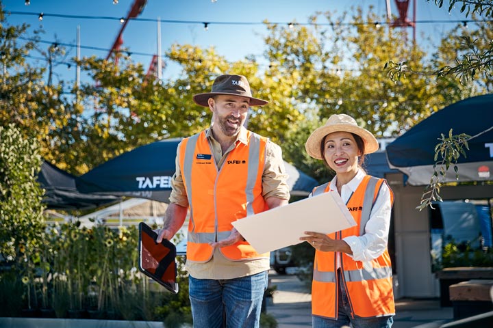 Two people wearing TAFE-branded high-visibility vests and hats, discussing outdoors. One holds a tablet, while the other holds a document folder.