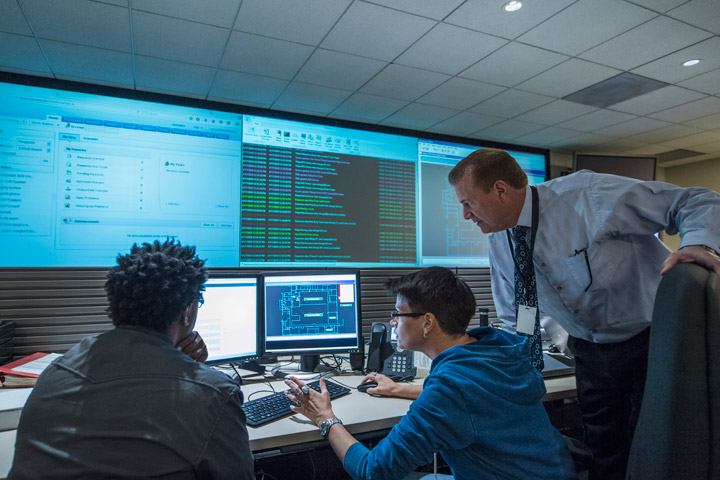 Acyber defence classroom with a teacher, two students on computers, and a large screen showing course data.