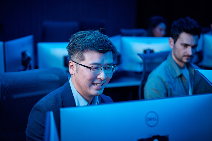 A TAFE NSW student engaged in a cyber defence class, smiling behind a computer screen. Other students do the same around him.
