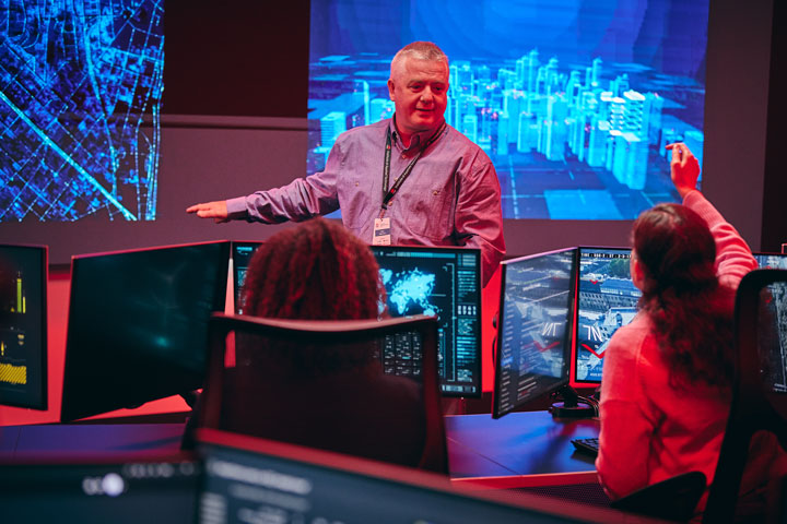 A TAFE teacher lectures on cyber attacks to engaged students with multiple screens at desks. Glowing cybersecurity projections enhance the learning environment.