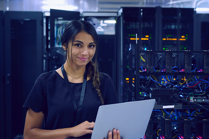 A young woman studying information and communications technology, holding an open laptop in front of a row of network servers.