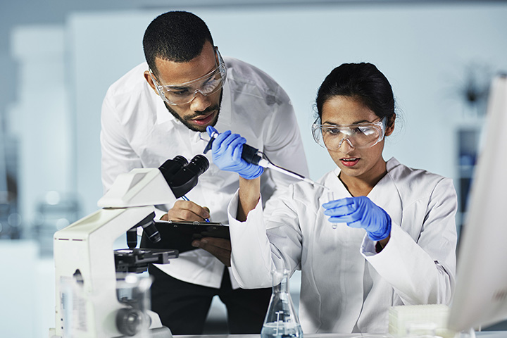 Two laboratory science students wearing lab coats and safety glasses, working with beakers and other lab equipment in a laboratory setting.