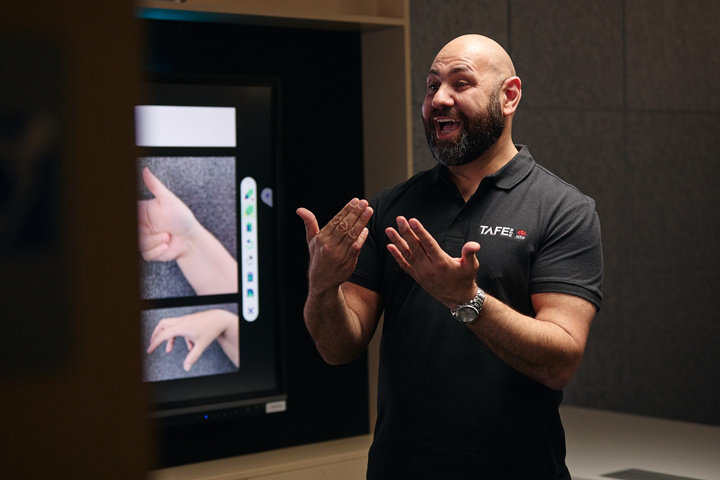 A man with a beard wearing a ɫֱpolo shirt is using sign language while standing in front of a screen displaying images of hands making different signs. The man appears to be engaged and expressive as he communicates.