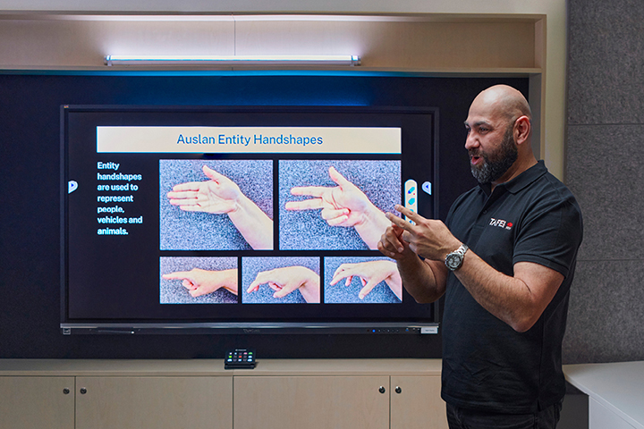 Male teacher teaching Auslan, gesturing while a screen behind him shows a range of different Auslan handshapes and signs.