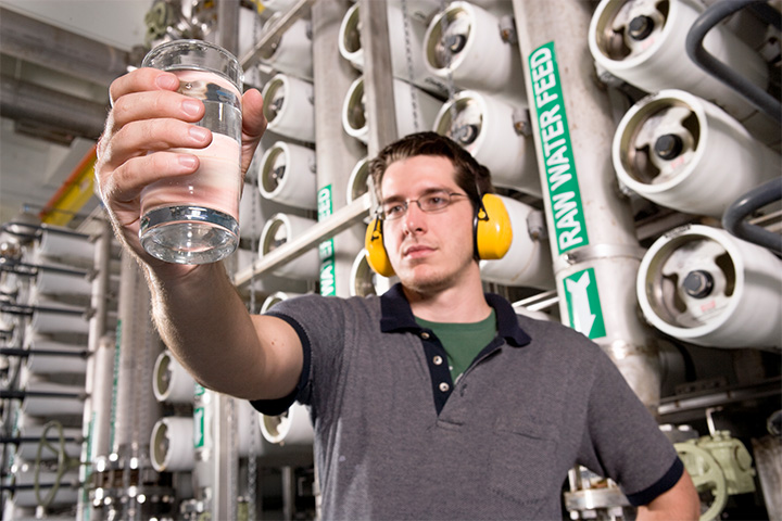 A young male mining engineer wearing safety glasses and ear protection inspects a beaker of liquid in a brightly lit factory environment. Industrial equipment and pipes are visible in the background.