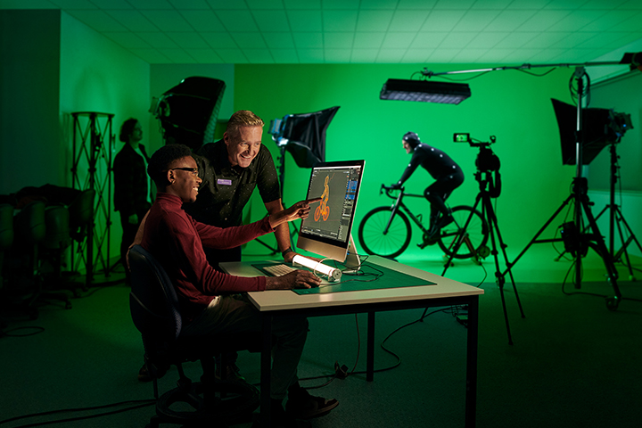Two people are smiling and looking at a computer screen showing an image of a man on a bike. In the background there is a green studio set up and a man is riding a bike. The image on the screen reflects this scenario.
