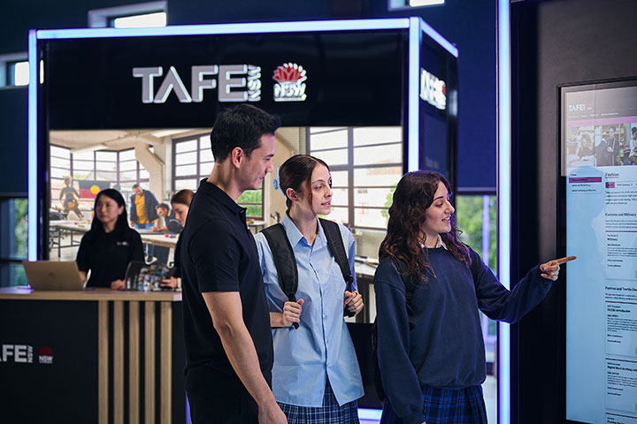Two young students and a career advisor at a TAFE NSW stand during a school expo, reviewing a display about available courses.