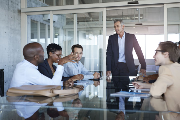A group of professionals gather around an office table. They are engaging in conversation and listening attentively. The office is bright and modern.