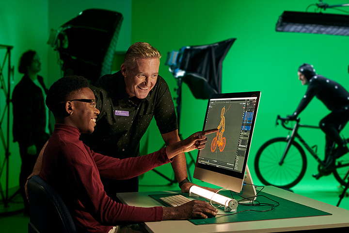 A young male designer works on a graphic design project on his computer in a brightly lit design studio. His teacher stands beside him offering guidance. In the background, a photographer shoots a scene involving a man riding a bicycle in front of a green screen.