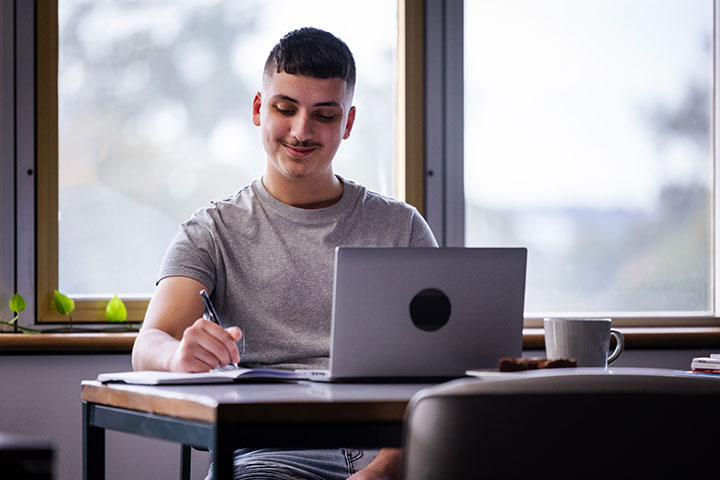 A young man smiles as he studies at his desk, writing in a notebook with his laptop open.