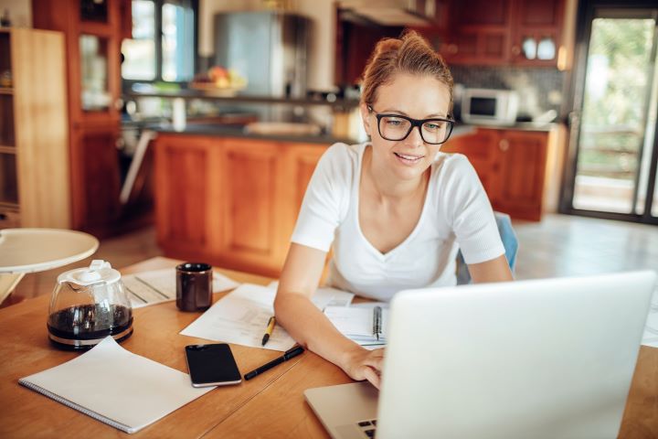 Student with black-framed glasses and a white T-shirt works on a laptop at her dining table. The table is cluttered with notepads, loose papers, pens, a smartphone and a cup of coffee.