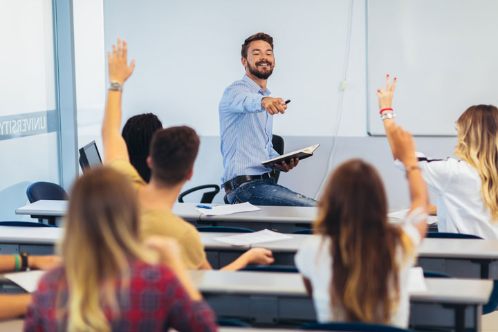 A smiling teacher sits at the front of his classroom engaging with students who are seated. He is pointing to a student with a raised hand indicating that she may answer a question. Several other students also have their hands up. The setting is bright and the students seem engaged in the discussion.