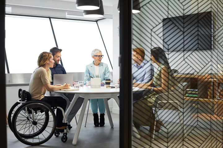 A diverse group of learners, including a woman in a wheelchair, discuss at a round table in a modern, architecturally designed space with emphasis on accessibility.