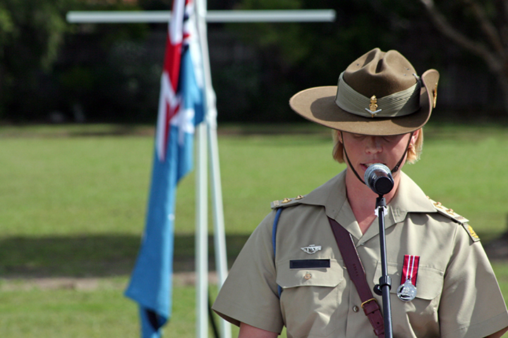 Australian military spokesperson in full official uniform speaking to a crowd using a microphone, with an Australian flag displayed in the background.