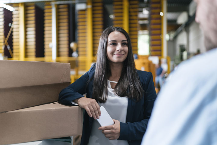 A woman in business attire stands in a warehouse setting, holding a smartphone and leaning on a stack of cardboard boxes. The background features industrial shelving and blurred activity, creating a professional and dynamic atmosphere.