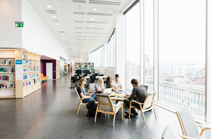 A group of students collaborate in a spacious and contemporary breakout area within a large open-plan library/study space. It features high ceilings and floor-to-ceiling windows that provide ample natural light with a view across a city-scape.