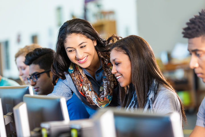 A young woman smiles as she assists another woman working on a computer. Both are seated in a computer lab, surrounded by other students focused on their screens. The atmosphere is collaborative and supportive.