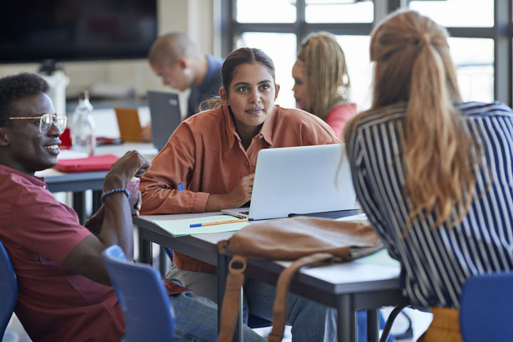 A group of students are seated around a table in a collaborative learning environment. One student is attentively listening to a peer while working on a laptop. Other students are engaged in their own tasks in the background. The scene captures an active and diverse study session, emphasising teamwork and focused learning.