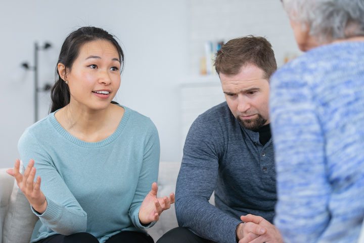 A woman in a light blue sweater gestures while speaking, sitting beside a man who appears deep in thought. They are engaged in a conversation with an older woman whose back is to the camera.