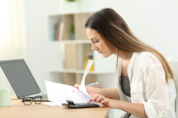 The image shows a woman sitting at a desk, focused on paperwork in front of her. She is holding a document and using a calculator. On the desk, there is also a laptop, a pair of glasses, a mug, and some additional papers. The setting appears bright and organised.