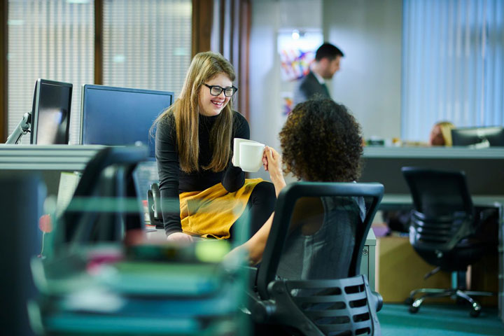 Two female students clinking mugs and smiling in an office with other students working in the background.