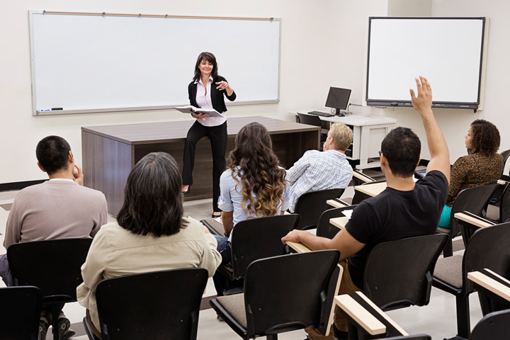 Students seated in rows actively participate in a classroom session, with one student raising a hand while the teacher stands at the front near a whiteboard, gesturing and holding papers during the lesson.