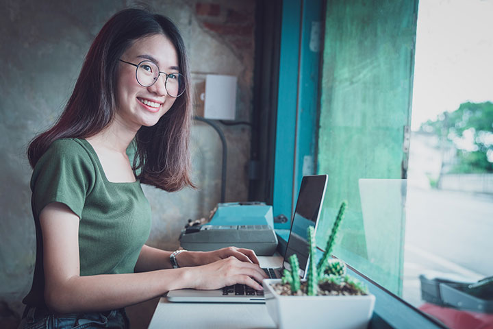 Woman smiling while using her laptop at a desk facing a street-facing window, with a cactus in the foreground.