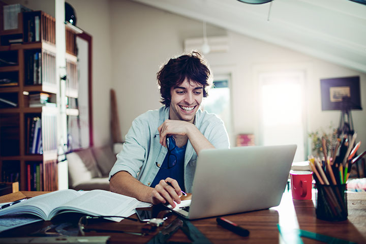 A young man with dark, shaggy hair is working at a desk on his laptop. He has an open book, stationery and coffee cup beside him. He is holding his glasses in one hand and appears to be in a home office setting with warm, natural light coming through a window in the background.