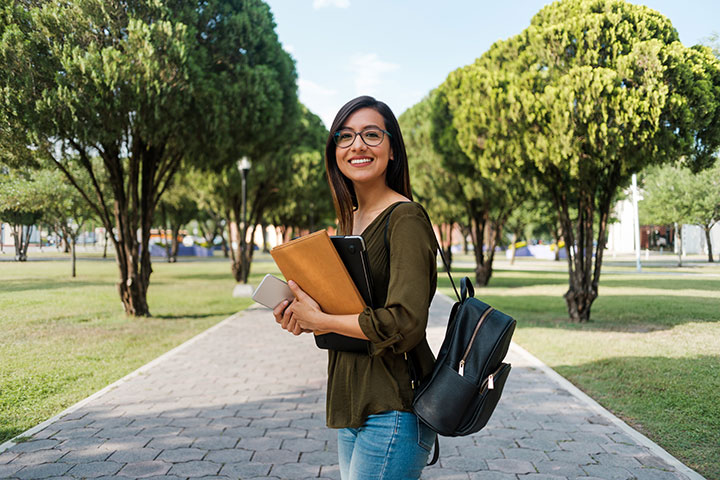 A young woman with glasses, wearing a green blouse and jeans, stands on a tree-lined path holding a folder, laptop, and smartphone, smiling confidently with a backpack over her shoulder.
