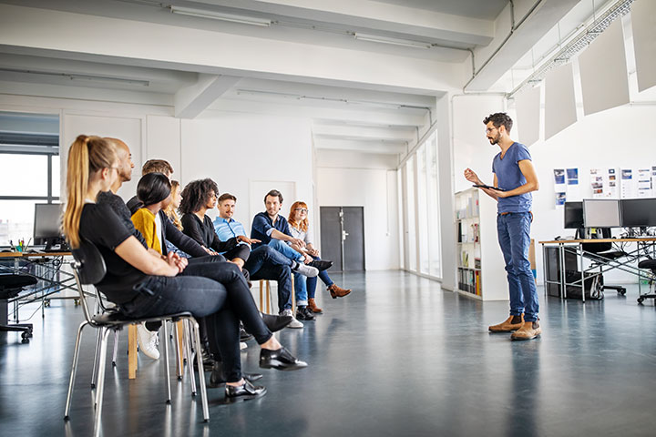 Man holding a tablet leads a discussion with a group seated in a semi-circle in a spacious, modern office with large windows.