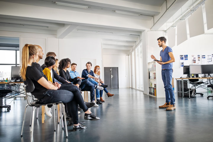 A group of people are seated in a semi-circle in a bright, modern office space. They are listening to a man standing in front of them. The man is holding a tablet and appears to be leading a discussion or presentation. 