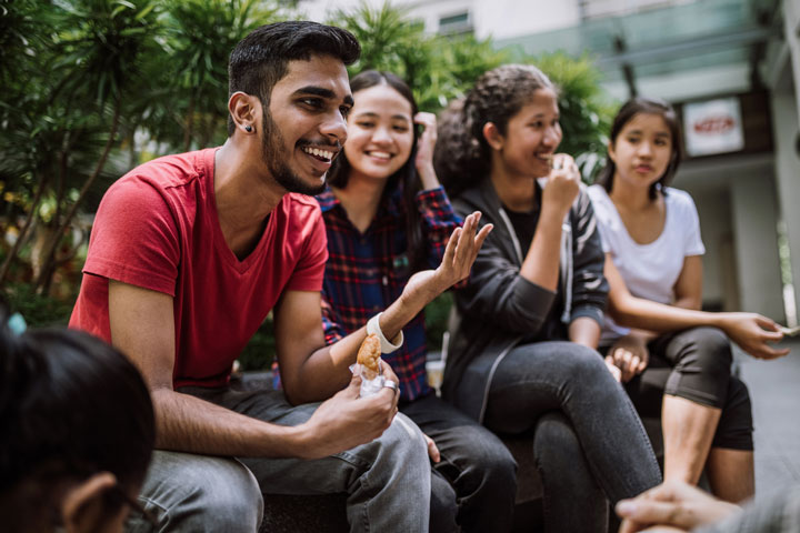 Group of young diverse people sitting outdoors, chatting and laughing together. The person in the foreground is wearing a red shirt and gesturing with their hand, while the others listen and smile. The background features green foliage and a modern building.